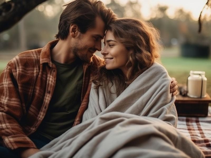 A couple snuggles under a blanket at a picnic under a tree.