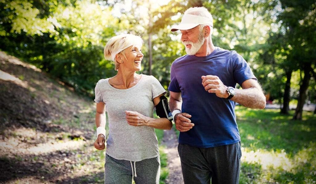 Two elderly people, a man and a woman, jogging together in a sunny park, smiling and conversing.