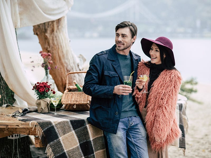 A couple toasting drinks at a picnic by a river, with a bridge in the background. the woman wears a hat and fluffy jacket; the man a blue jacket.