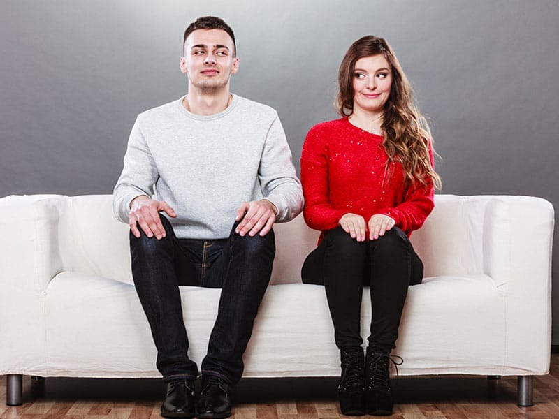 Two people sitting side by side on a white sofa, with the man wearing a gray sweater and jeans and the woman wearing a red glittery top and black pants. they both have a slight smile and are looking towards the camera.