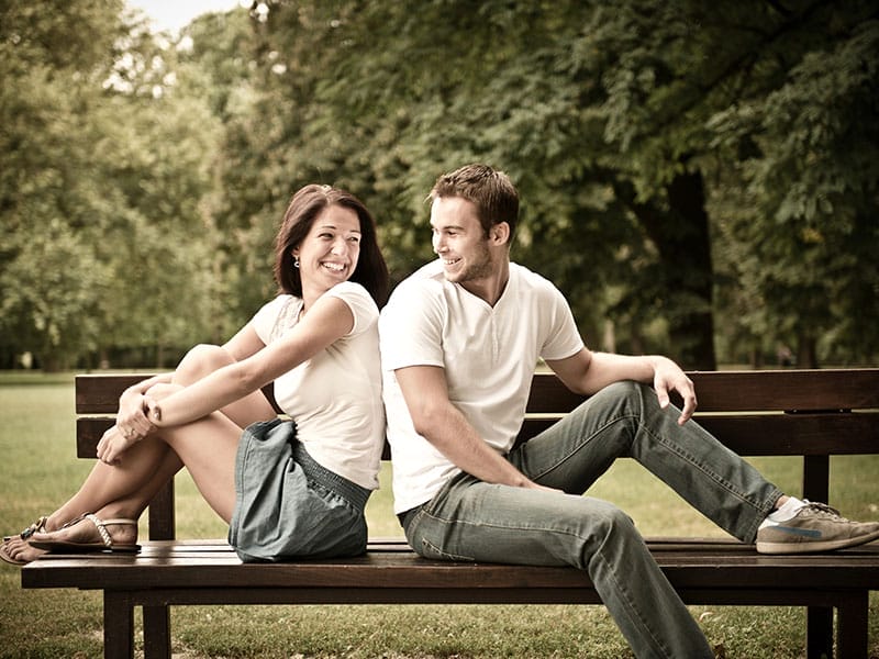 A man and a woman sitting back-to-back on a park bench, smiling at each other, surrounded by trees.