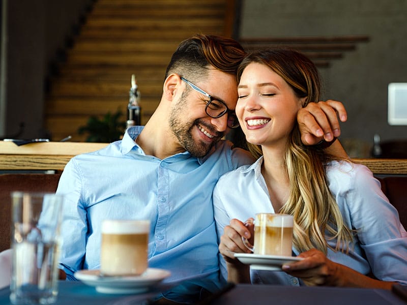 A happy couple, a man and a woman, laughing and enjoying coffee together in a cozy cafe setting.
