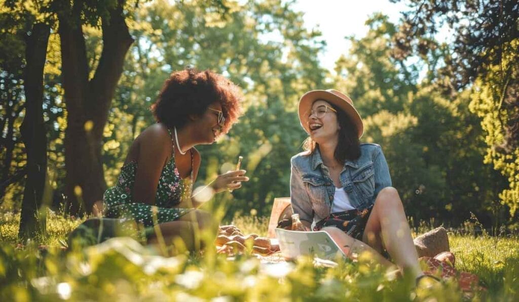 Two women laughing together during a picnic in a sunny park, sitting on the grass with a laptop and snacks around them.