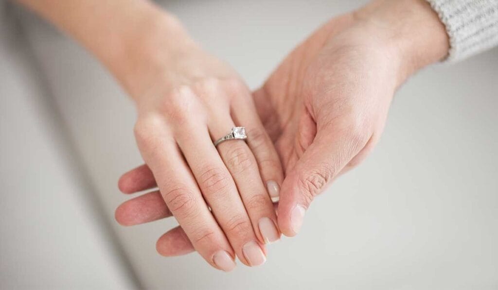 Two hands gently hold a left hand, which wears a Princess Cut engagement ring.