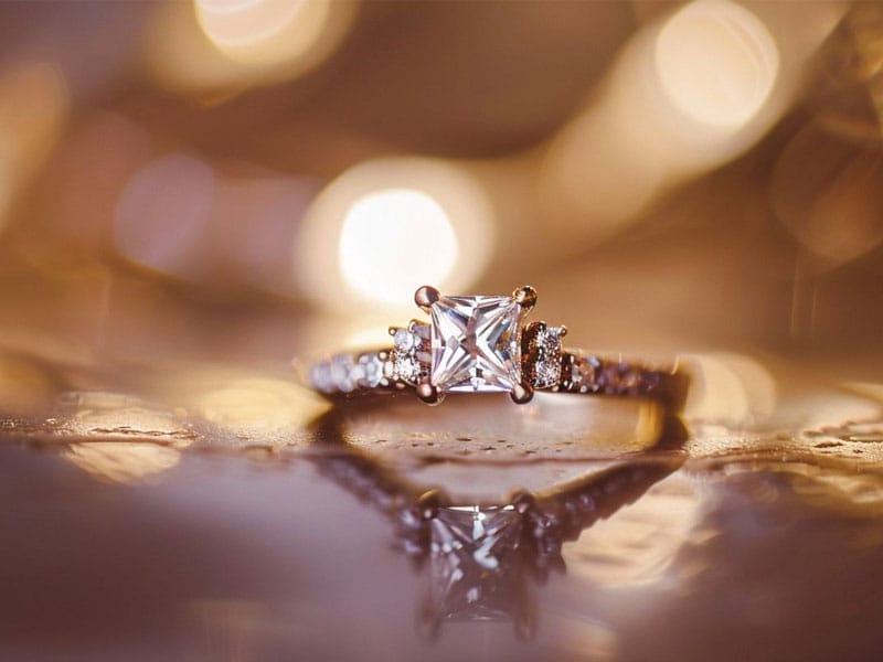 Close-up of an engagement ring with a square-cut central diamond and smaller diamonds on the band, set against a warm, blurred background with bokeh lighting.