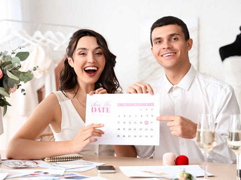 A smiling woman and man hold up a "Save the Date" calendar for their wedding, pointing to a specific date. Various wedding planning items are on the table in front of them.