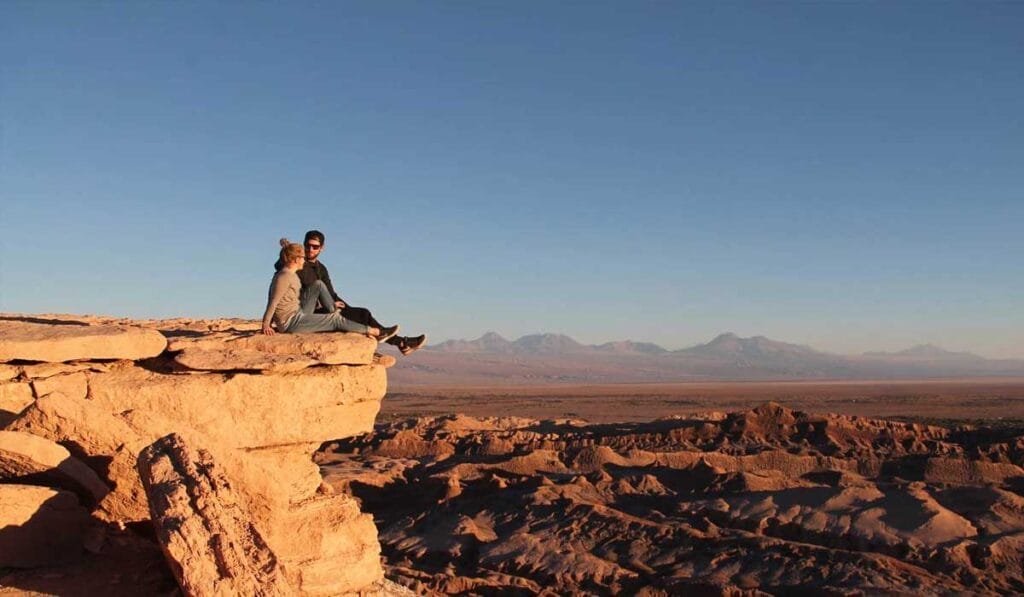 Two people sit on the edge of a high cliff overlooking a vast desert landscape under a clear blue sky.