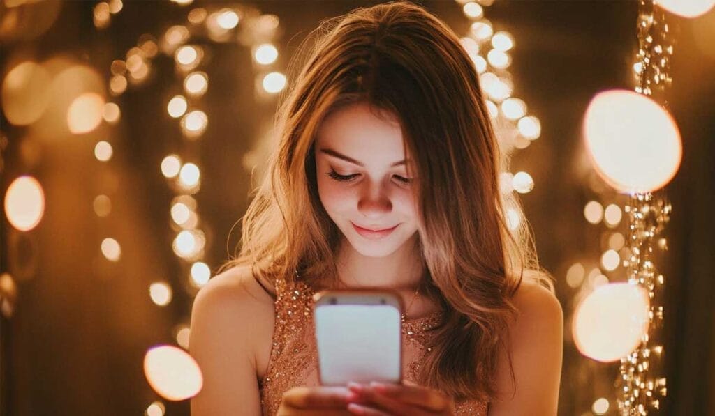 A young woman looks down at her smartphone while surrounded by warm, glowing string lights. She is smiling slightly.