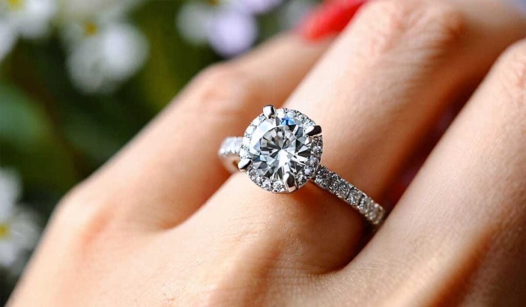 Close-up of a hand wearing a silver engagement ring with a large round diamond and smaller diamonds embedded on the band, set against a blurred floral background.