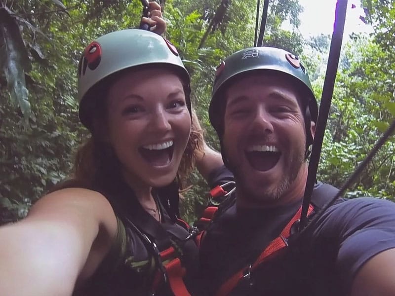 Two people wearing helmets and harnesses are smiling widely while ziplining in a forested area.