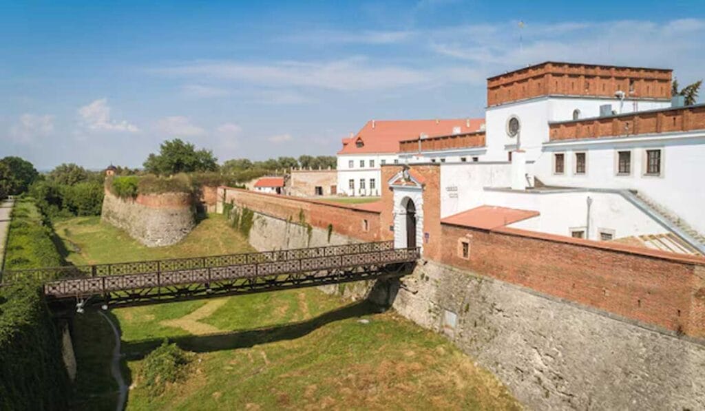 A historic fortress with white buildings, red roofs, and a wooden bridge over a moat on a sunny day.