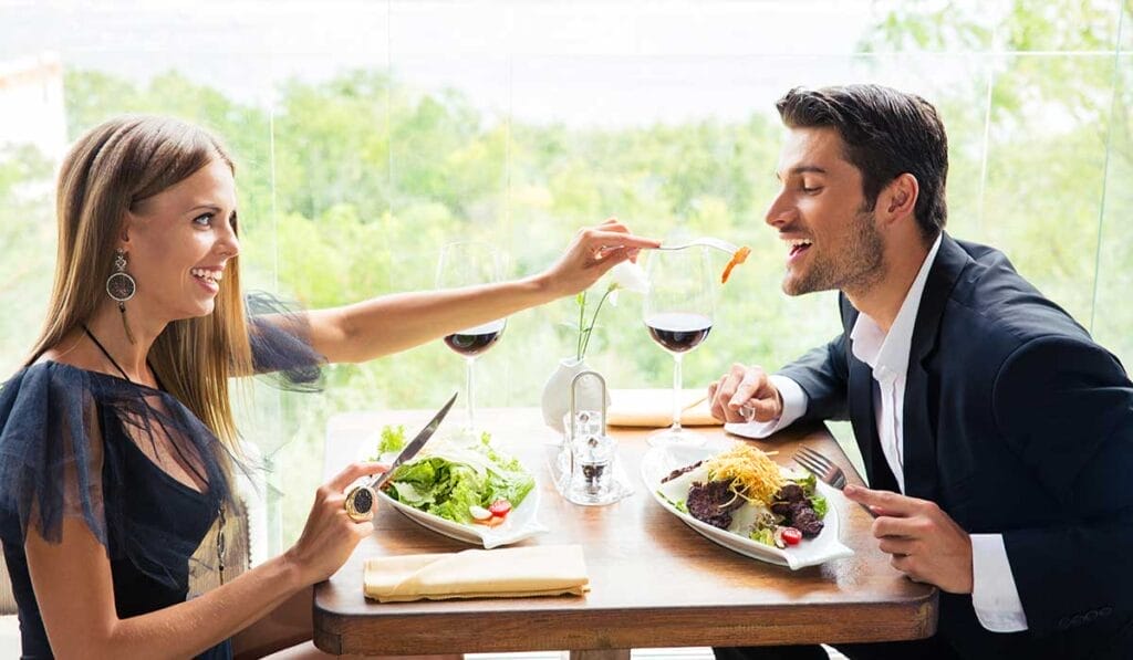 A woman and a man are seated at a restaurant table, enjoying a meal. The woman is feeding pasta to the man. Both are smiling and have glasses of red wine in front of them.