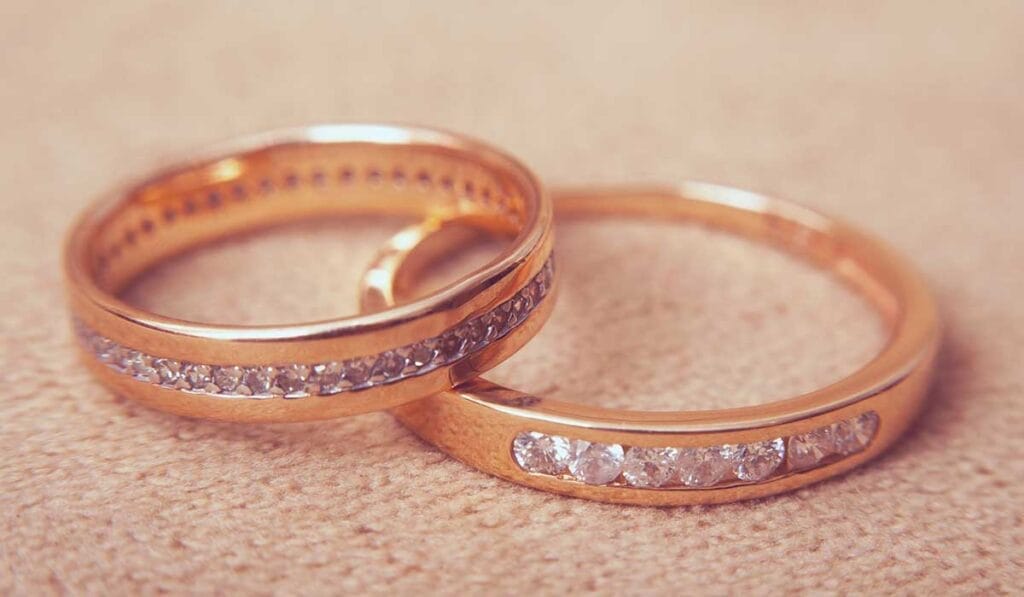 Two gold rings adorned with rows of small diamonds are lying on a beige fabric surface.