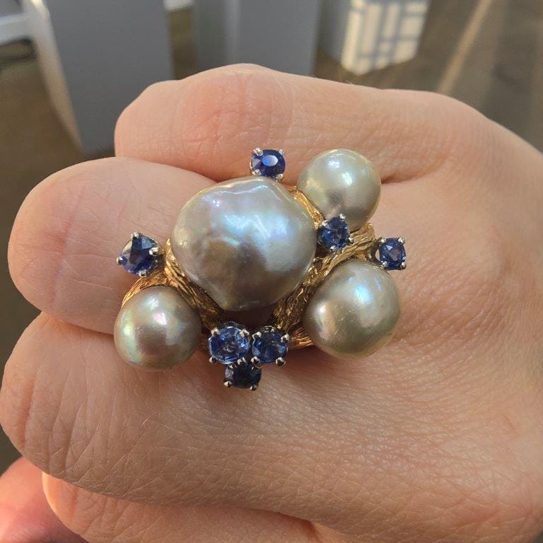 A ring with a large, irregular pearl at the center, flanked by three smaller pearls and seven small blue gemstones on a gold band, worn on a hand.
.