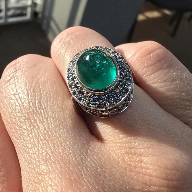 Close-up of a hand wearing a silver ring with a large green gemstone surrounded by smaller blue stones.