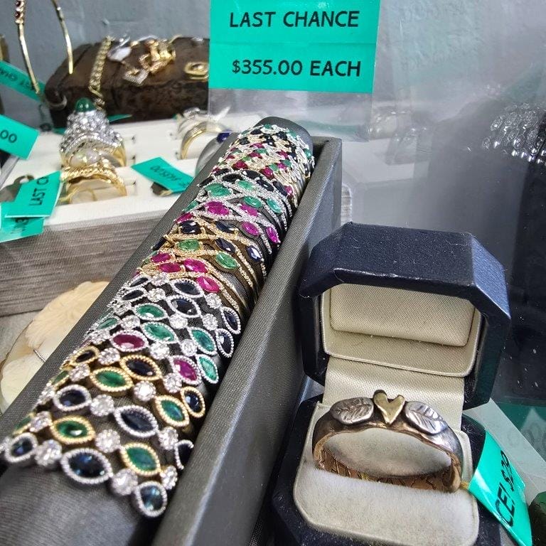 Display of various pieces of jewelry, including colorful bracelets and rings. A sign states, "LAST CHANCE $355.00 EACH." A ring with heart shapes is prominently placed in a box.