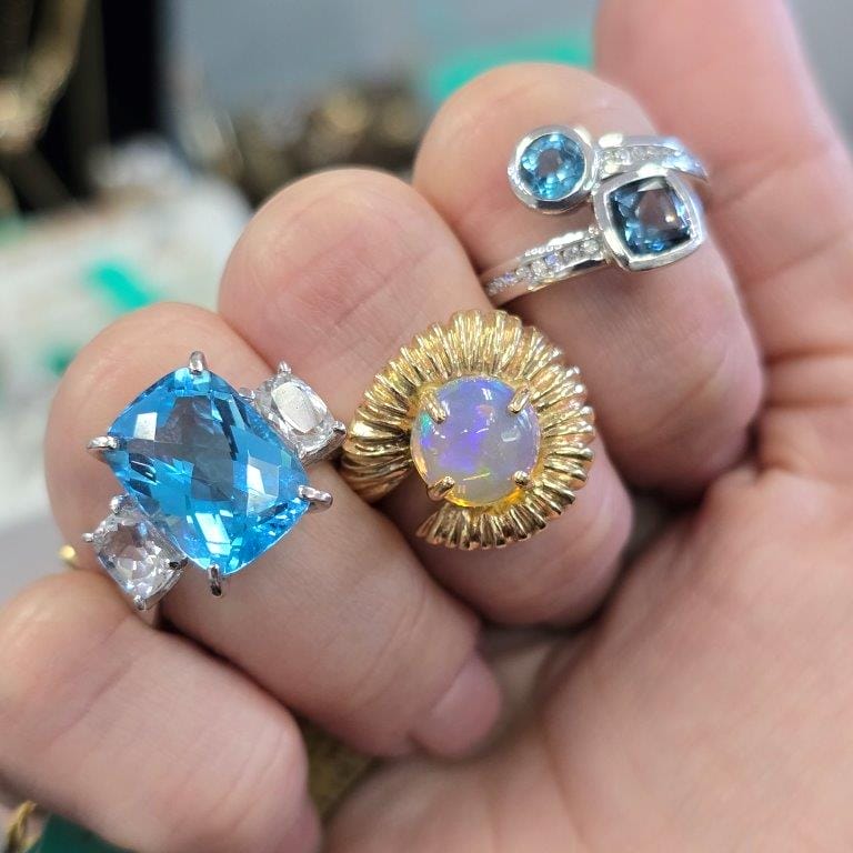 A hand displaying three rings: one with a large blue gemstone, one with a central opal set in a gold sunburst design, and one with a blue gemstone-adorned band with smaller blue gemstone.