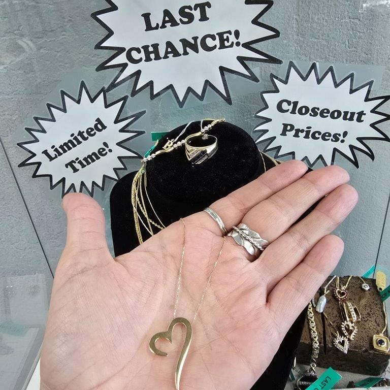 A hand holding a heart-shaped necklace in front of a display with signs reading "LAST CHANCE!", "Limited Time!", and "Closeout Prices!" showcasing various jewelry.