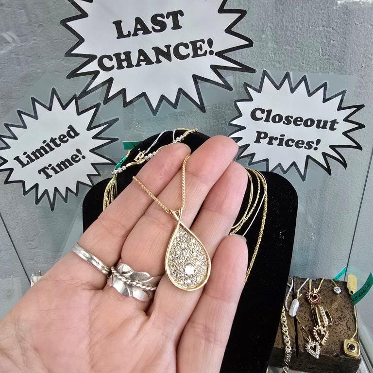 A hand holds a teardrop-shaped gold necklace with a diamond pattern in front of a display. Signs in the background read "LAST CHANCE!", "Limited Time!" and "Closeout Prices!.