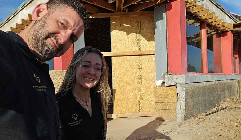 David and Aubrey stand in front of a partially constructed building with plywood covering the entrance. The man on the left and the woman on the right are smiling at the camera.