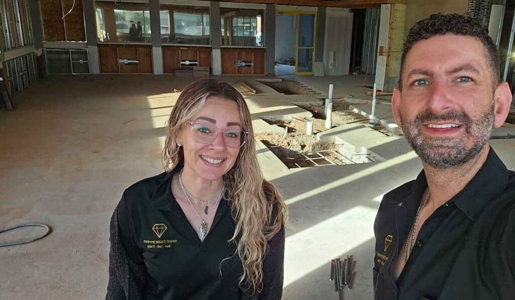 Two people in uniform shirts stand in a partially constructed building with exposed flooring and unfinished walls, both smiling at the camera.