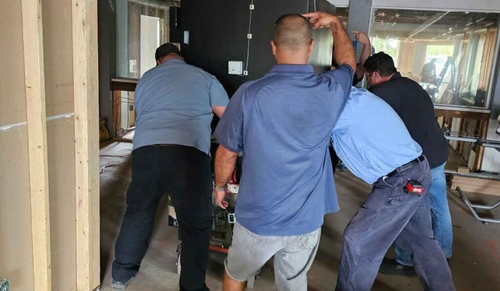 Four people are seen from behind, working together to move a large, dark object on a cart in an unfinished room.