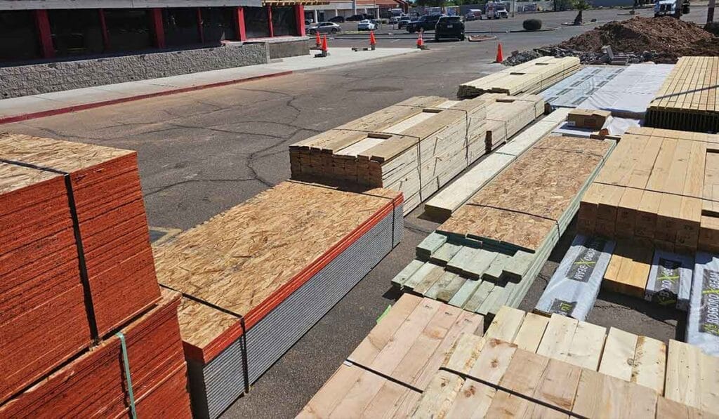 Stacks of lumber and building materials are organized on a paved area next to a storefront with construction cones in the background.