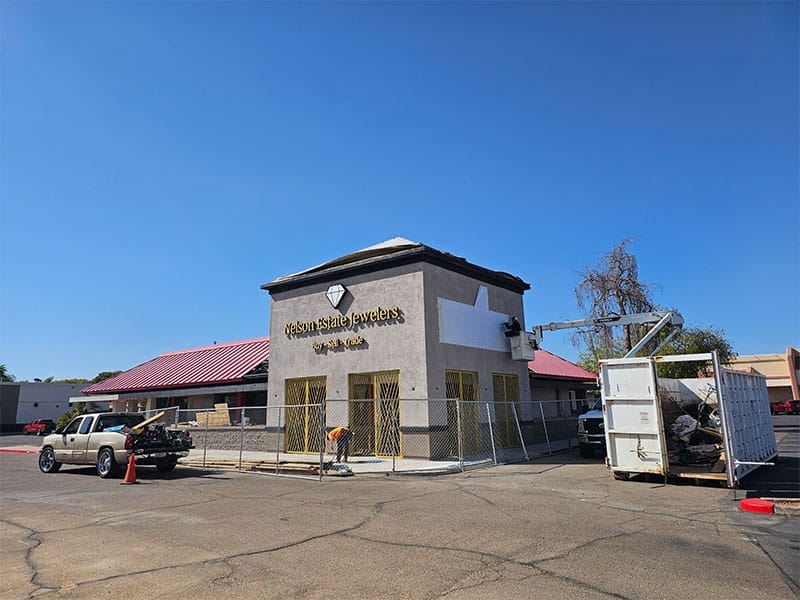 A worker on a lift removes signage from a jewelry store. A fenced-off area surrounds the building, and a truck and dumpster are parked nearby.