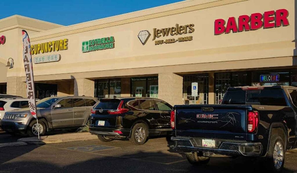 A strip mall with businesses including an acupuncture clinic, insurance agency, jewelry store, and barber shop. Several cars are parked in front.