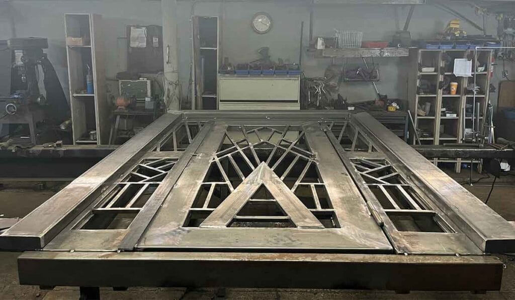 A large metallic gate with intricate geometric patterns lies flat on a workbench in a workshop, surrounded by various tools and equipment.