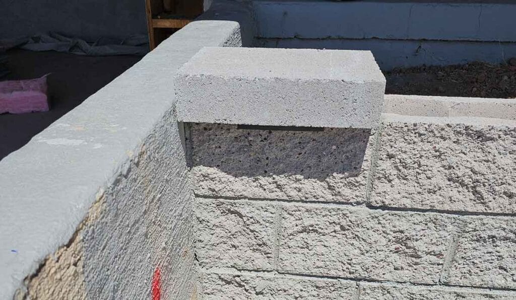 A single gray cinder block is partially set on top of a corner of a concrete block wall under construction. There is a hollow space beneath the elevated cinder block.