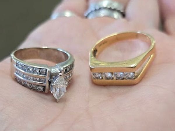 A hand displays two rings: a silver ring with a large marquise-cut stone and smaller stones on the band, and a gold ring with five small stones set in a row.