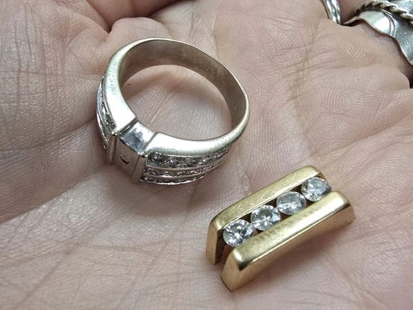 A hand holding a silver ring with an empty stone setting and a gold piece with three embedded clear stones.