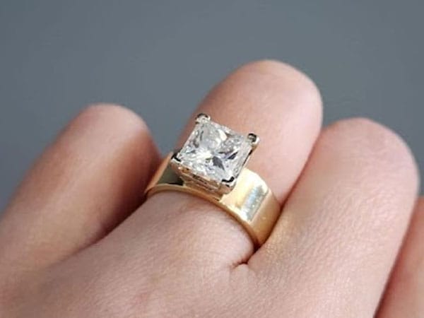 Gold ring with a large square-cut diamond on a person's finger against a gray background.