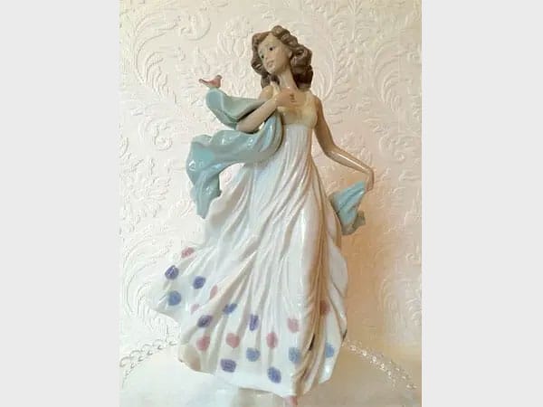 A porcelain lladro figurine of a woman in a flowing white dress with colorful floral accents, holding a light blue shawl.