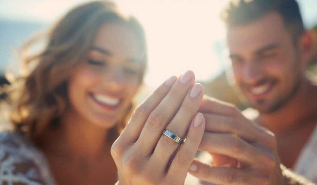 A smiling couple displays a silver ring in a sunlit setting.